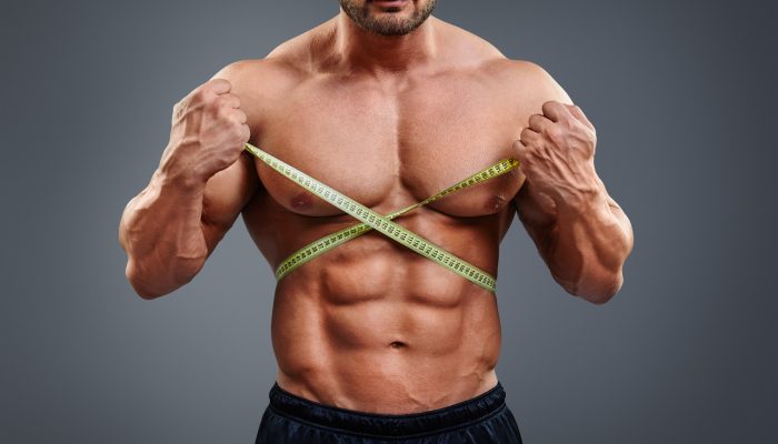 Personal fitness trainer holding tape measure isolated over gray background. Closeup of muscular man measuring his waist. Doing perfect six pack abs.
